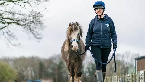 A Blue Cross equine handler leads a small grey pony along a gravel track. The handler is smiling and the ponies ears are pricked forward.